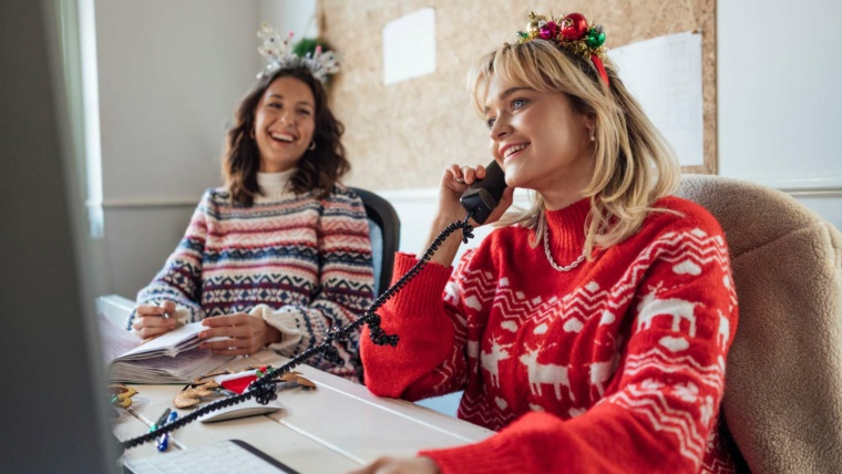 Two women in festive sweaters sit at an office desk; one is on the phone, and the other is writing notes.